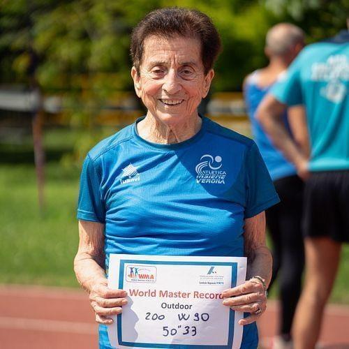 Emma Maria Mazzenga smiling on a track, holding a World Master Record sign for the women's 90+ 200 meters