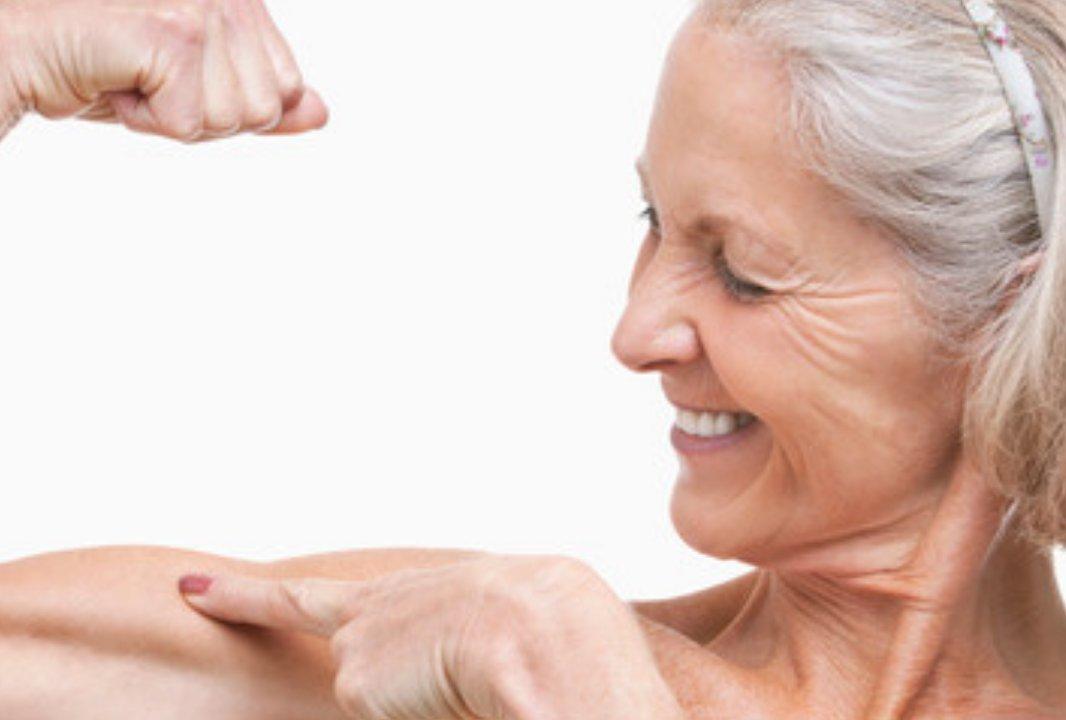 A smiling older woman flexing her arm and pointing to her bicep