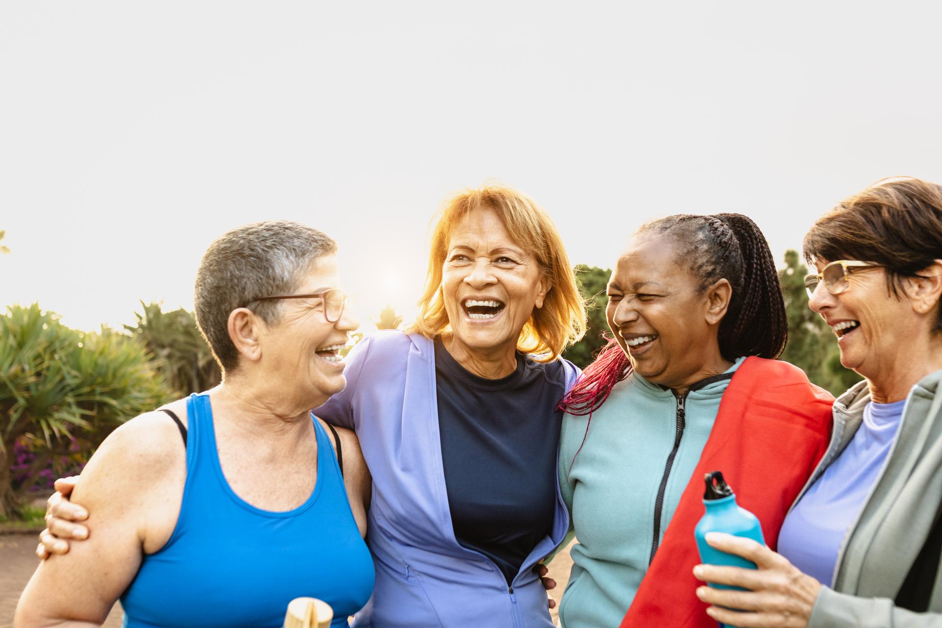 Four older women in workout clothes laughing arm in arm outdoors